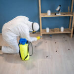 A professional in a white hazmat suit and respirator uses a specialized sprayer to demonstrate how do pest control get rid of cockroaches by treating a floor and wooden shelving unit in a home.