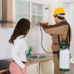 A professional technician providing 24 hour pest control services, spraying inside a kitchen cabinet while a homeowner watches.